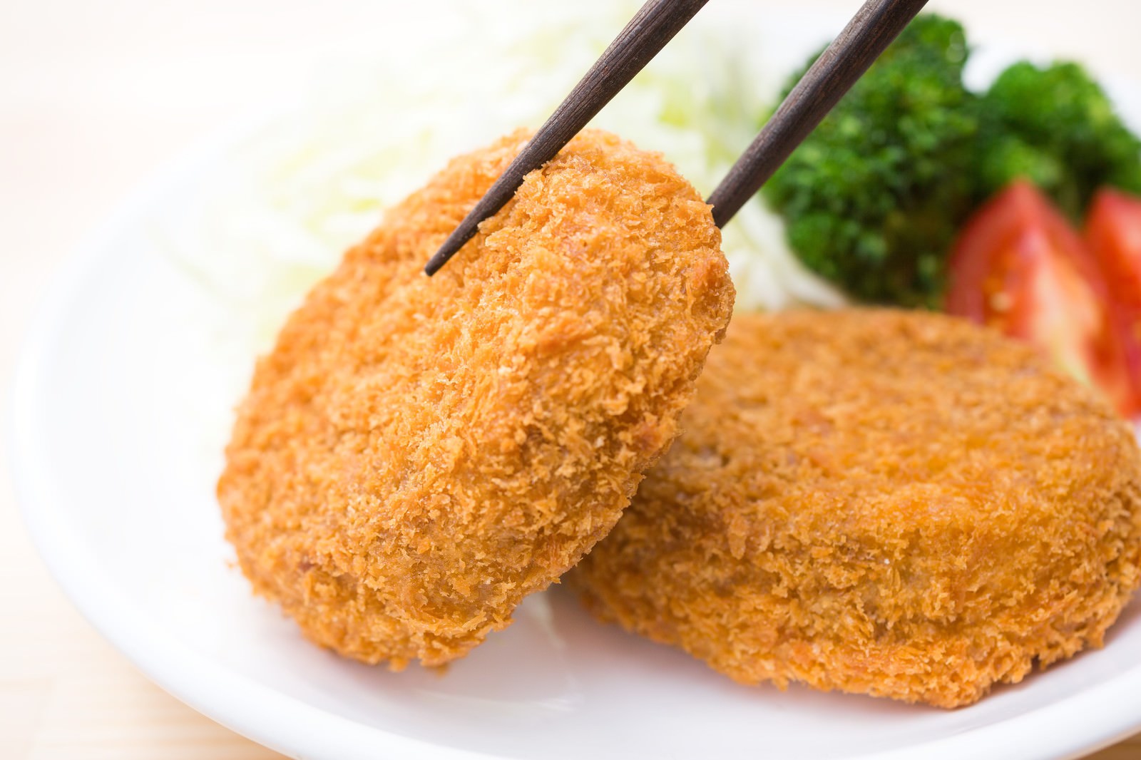 箸でつまんだ揚げたてのコロッケと白い皿に盛られた付け合わせ