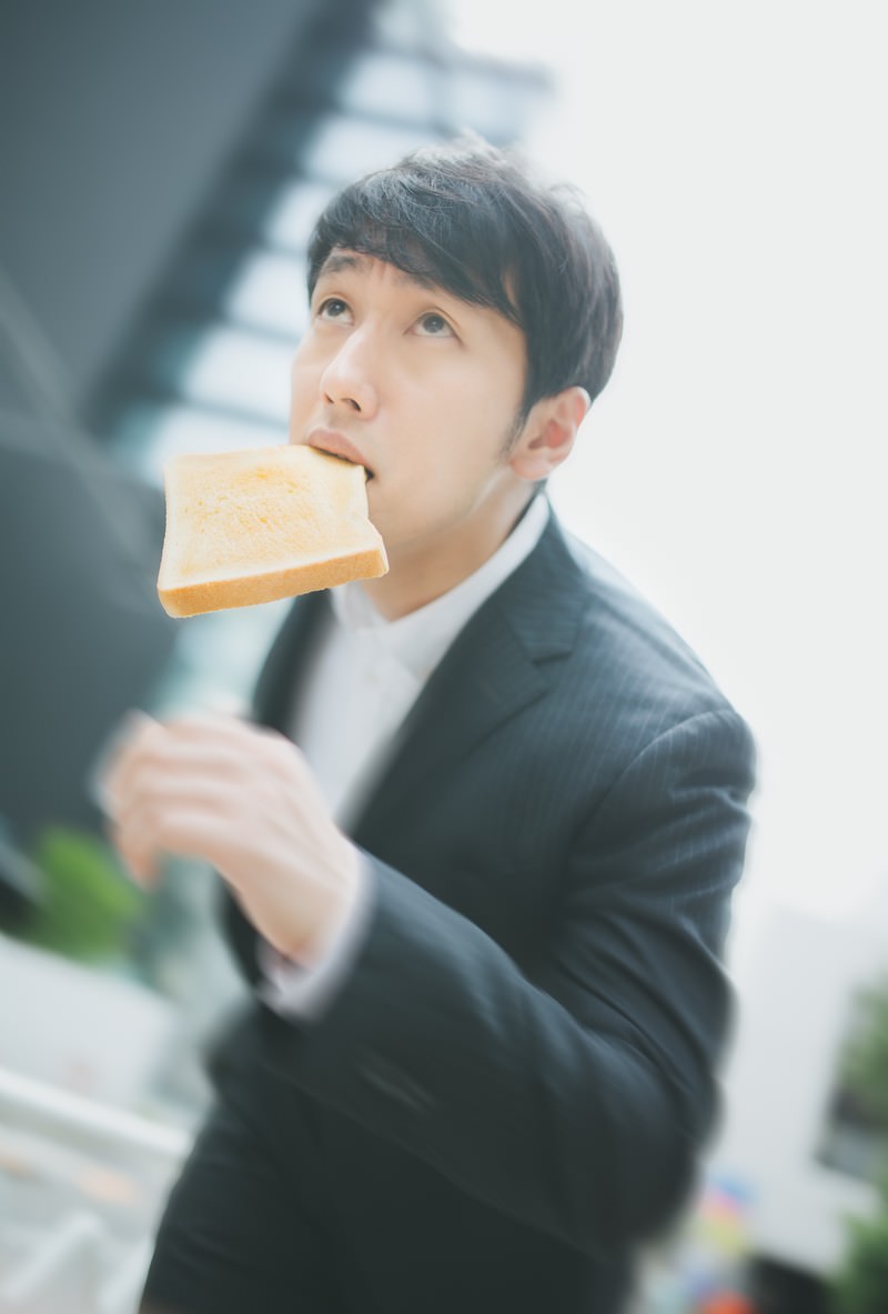 トーストを咥えて下から見上げた角度のスーツ姿のビジネスパーソン［モデル：大川竜弥］