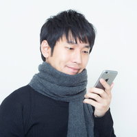 灰色のマフラーを巻きスマホを両手で見る黒い縦縞セーターの男性の写真