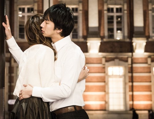 夜間の木造建築前で白いニットとシャツの男女が後ろ姿で抱き合う写真