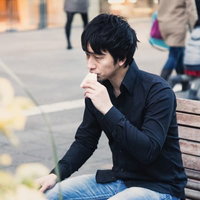 屋外のベンチに座り手を口元に当てる黒いシャツにジーンズの黒髪男性の写真