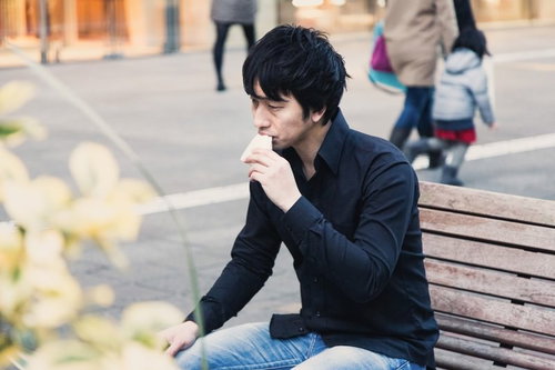 屋外のベンチに座り手を口元に当てる黒いシャツにジーンズの黒髪男性