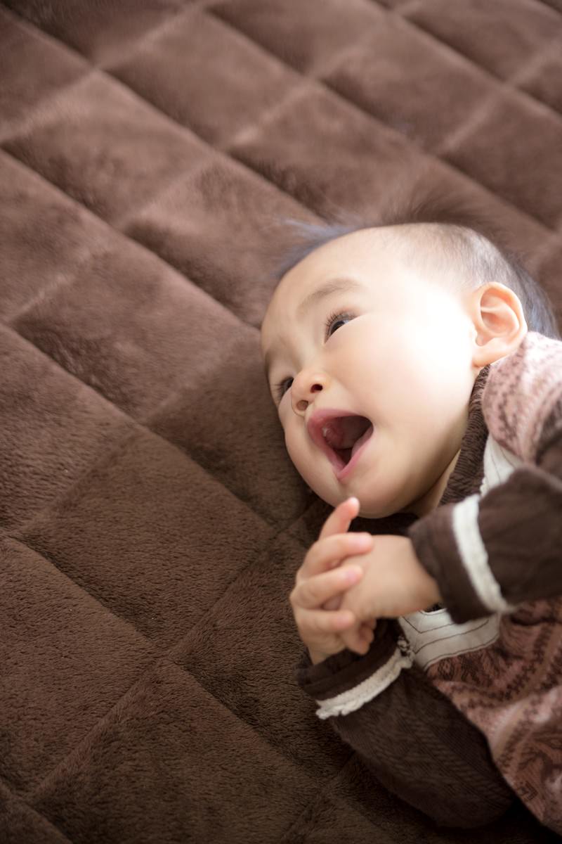 茶色のマットの上に仰向けに寝た赤ちゃんが口を大きく開けている［モデル：あんじゅ］