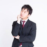 右手を顎に当て左腕を組んで考えるスーツの男性の写真