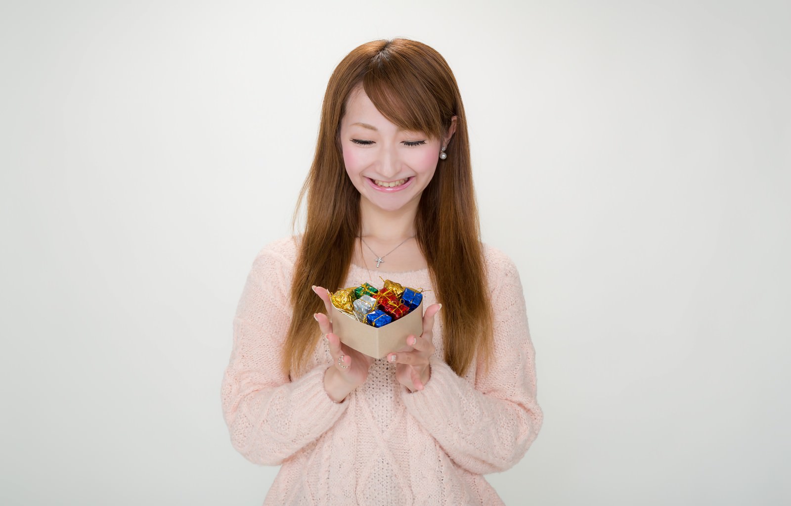 ベージュのギフトボックスをたくさんのリボンで飾られたプレゼントが詰まっており、ピンク色のカーディガンを着た女性が両手で持ち笑っている［モデル：to-ko］