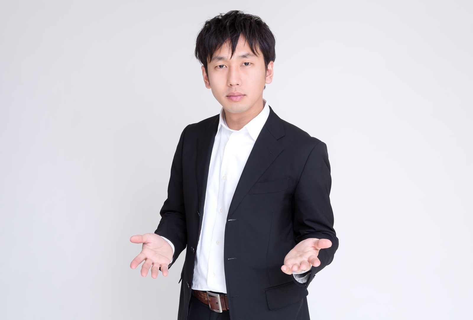 黒いジャケットと白いシャツの男性が白い背景の前で両手を広げて立っている［モデル：大川竜弥］