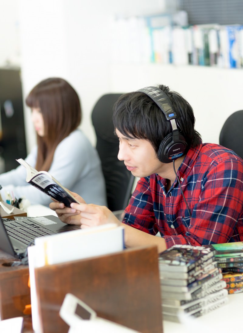 ヘッドフォンをつけてオフィスで漫画を読む赤いチェックシャツの男性