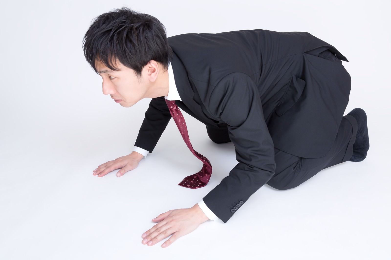 深紅色のネクタイを付けた黒いスーツ姿の男性が、両手を床につけて膝をつき、頭を下げた土下座の姿勢をしている［モデル：大川竜弥］