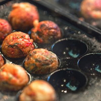 油が飛び散る黒い鉄板の丸い穴で焼かれる茶色のたこ焼きの写真