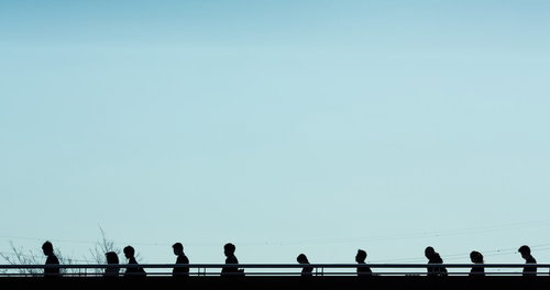 明るい空を背景に歩道橋を歩く複数の人のシルエットの横一列