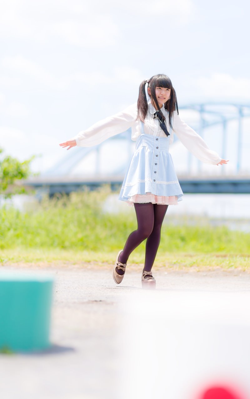 河川敷の舗装路でツインテールの女の子が水色のジャンパースカートで両腕を広げている［モデル：こころ］