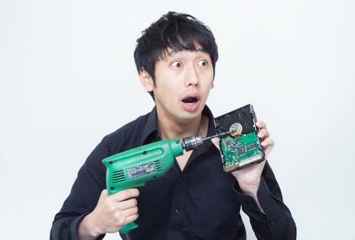 ドリルとHDDを持って焦った表情で目を見開く黒シャツの男性