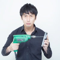 緑色の電動ドリルとスマホを持って正面を向く黒シャツの男性の写真