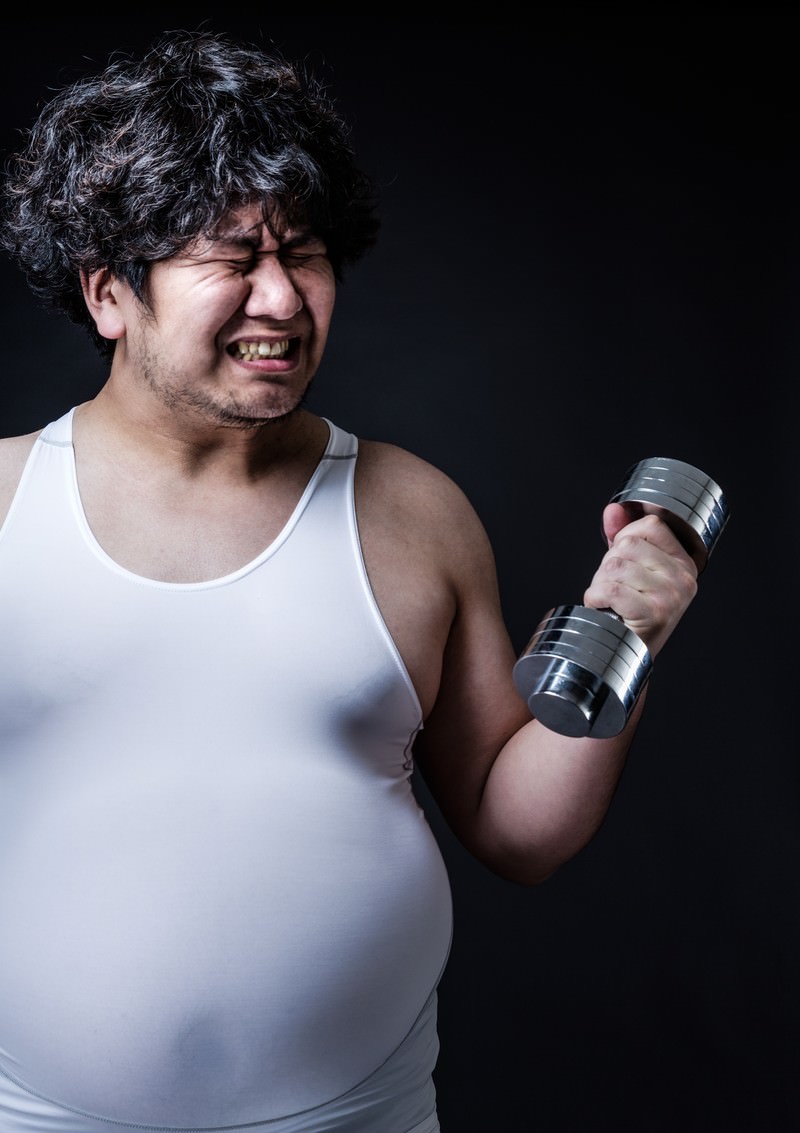 ダンベルを持ち苦しい表情でトレーニングする肥満男性［モデル：あまのじゃく］