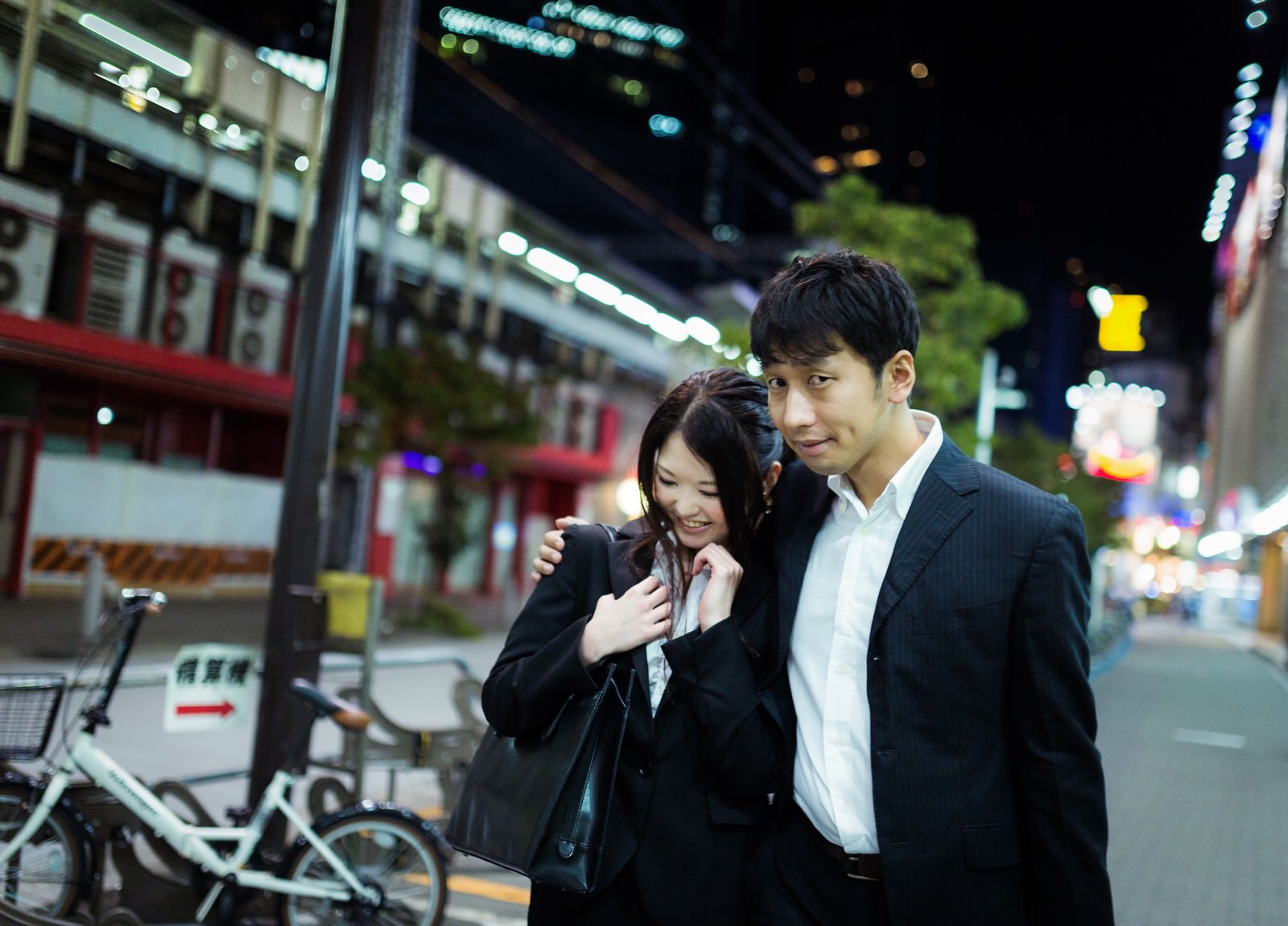 スーツ姿の男性が女性の肩に腕を回し、夜の繁華街で寄り添っている場面。背後はネオンサインと店舗灯［モデル：大川竜弥 シマヅ］