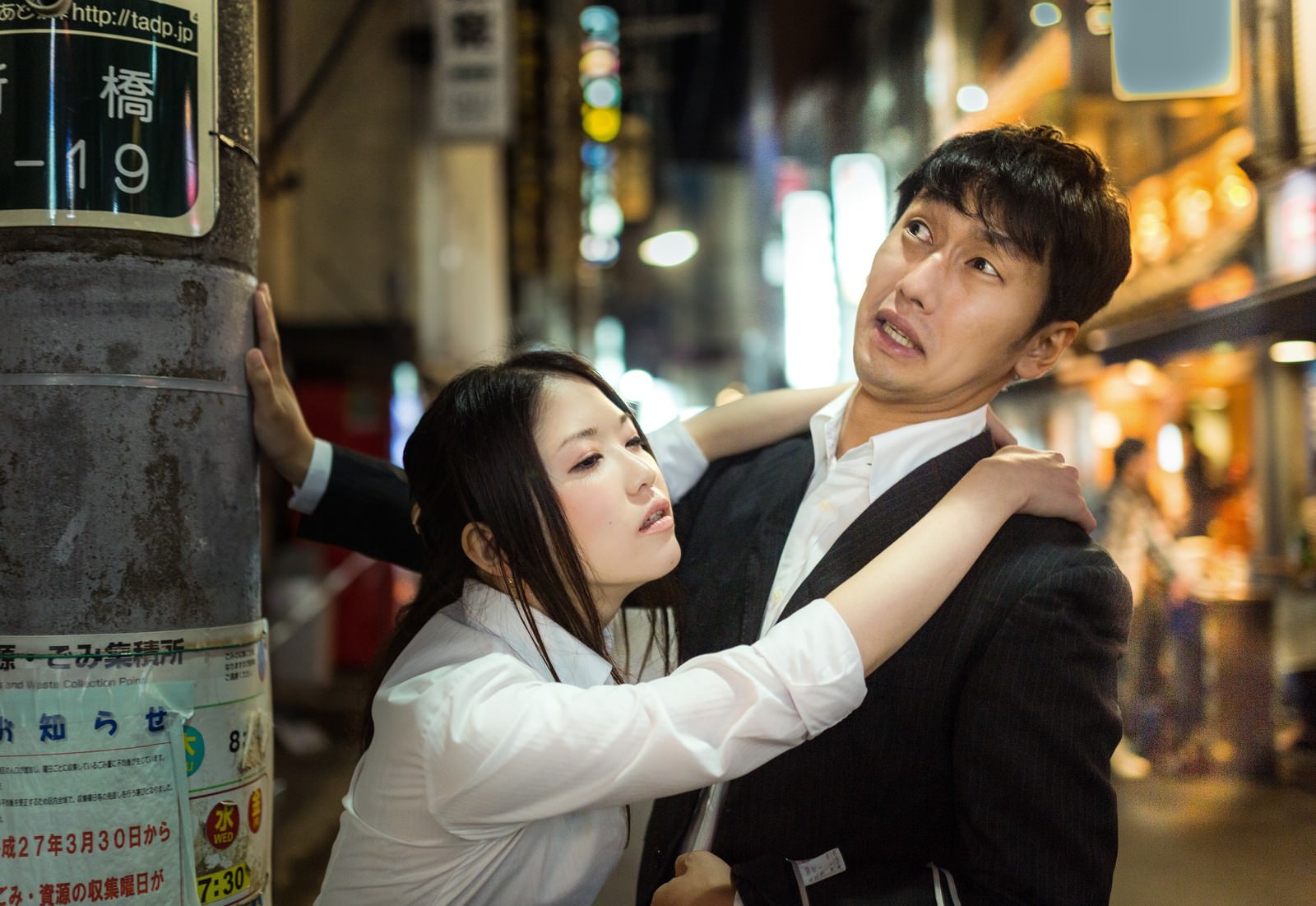 夜の繁華街で電柱に寄りかかり上を見上げる白シャツの女性と男性［モデル：大川竜弥 シマヅ］