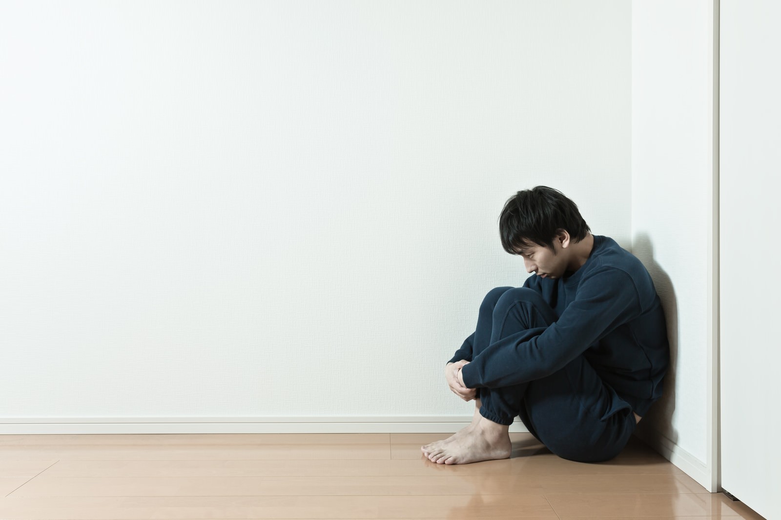 白い壁の室内で青緑色の服の男性が木の床に座り膝を抱えてうつむいている［モデル：大川竜弥］