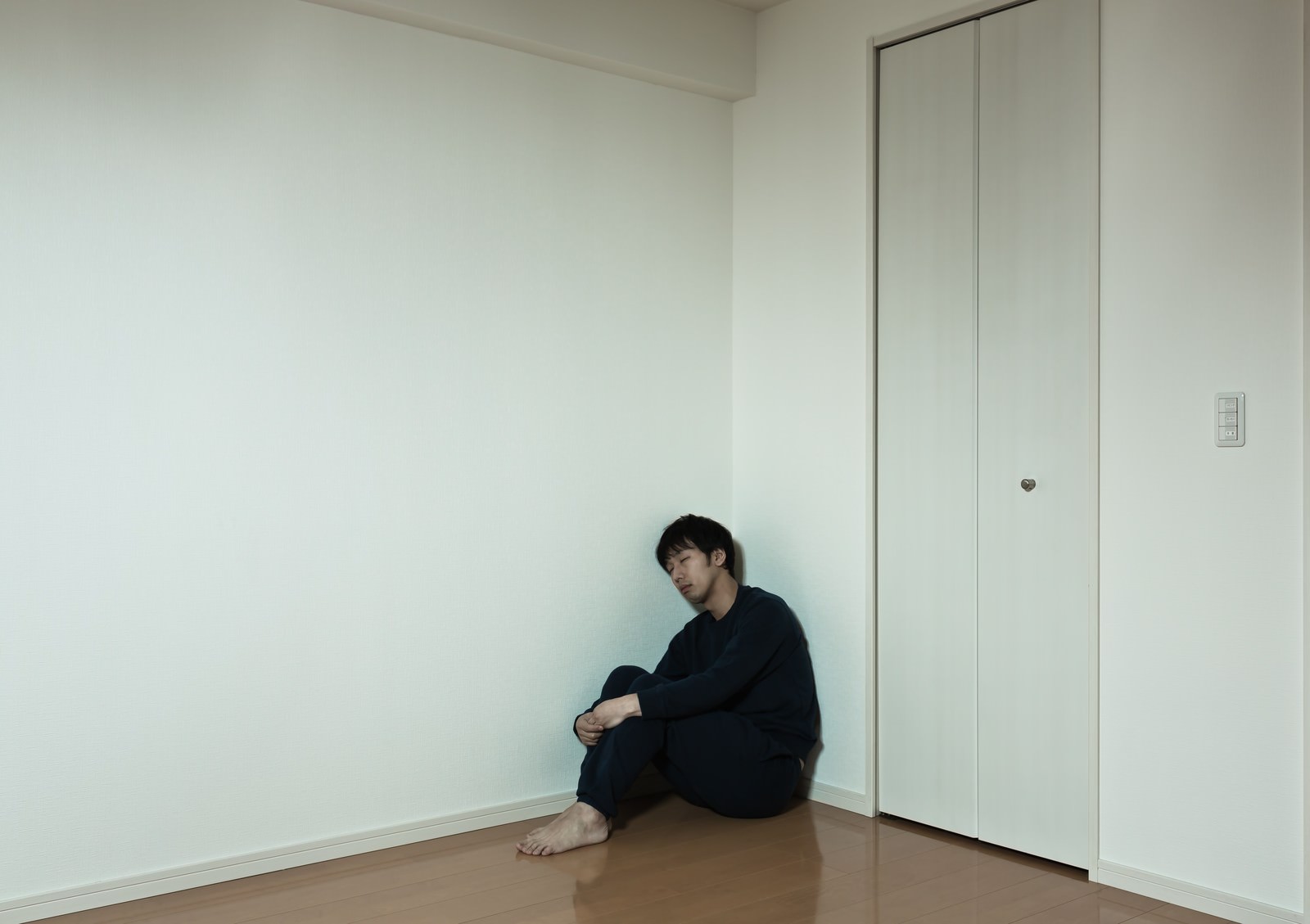 白い壁と茶色の床の部屋の隅で黒い服の男性が床に座り込んでいる［モデル：大川竜弥］