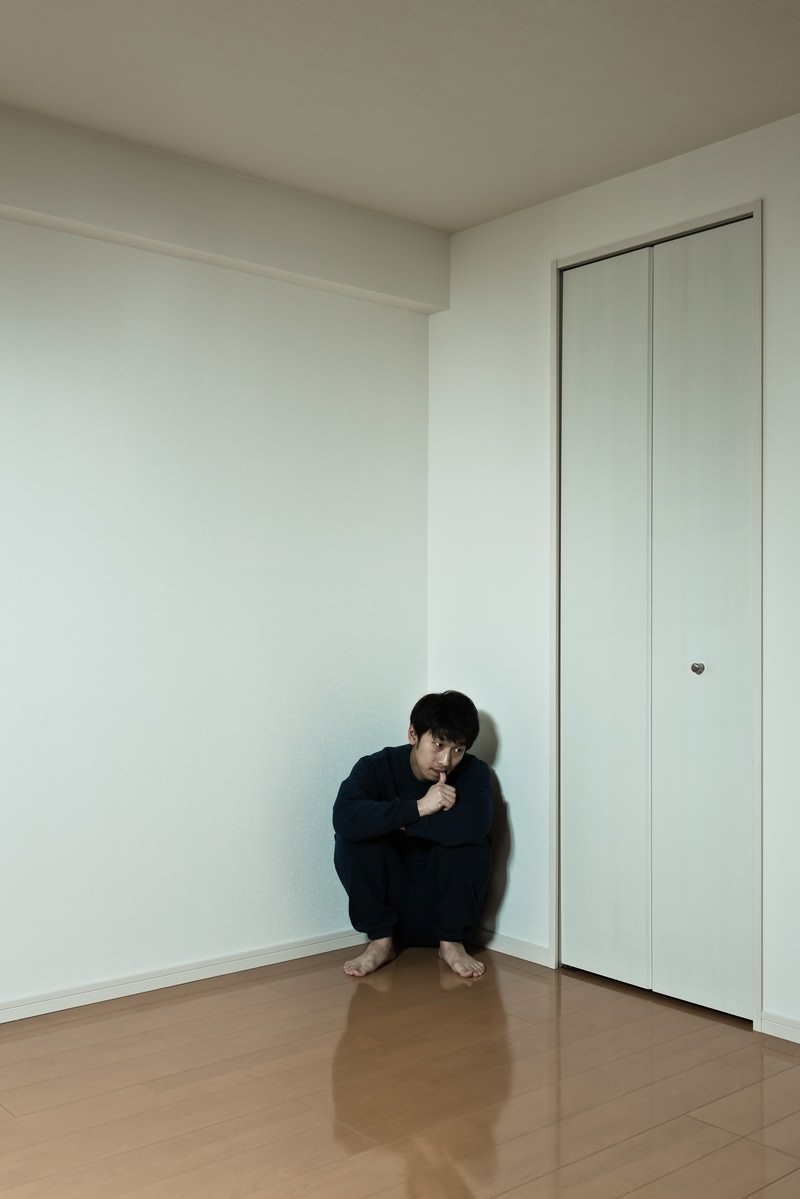 白い壁の部屋の隅で黒い服の男性がしゃがみ込んで膝を抱えている［モデル：大川竜弥］
