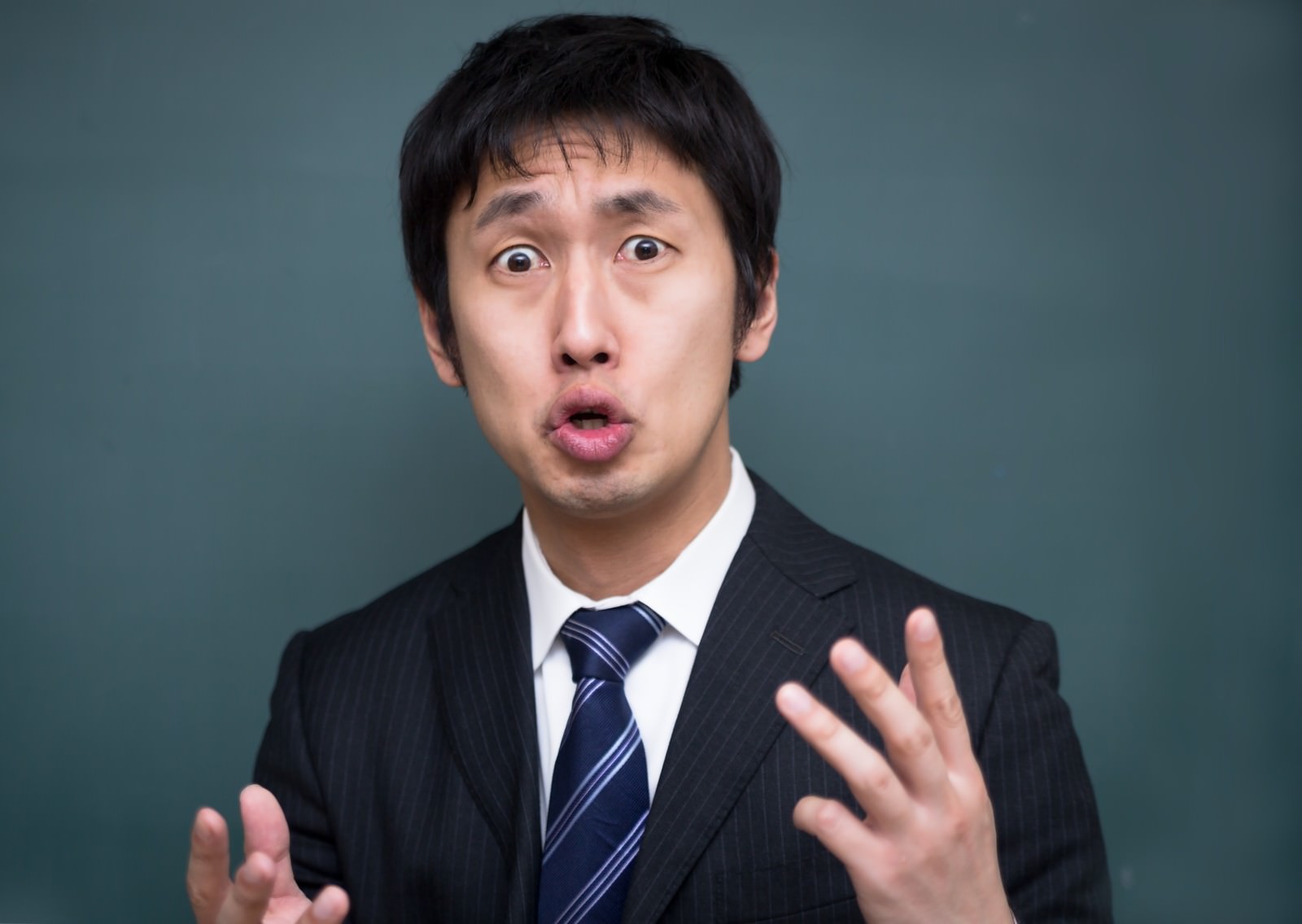 紺ネクタイ・スーツ姿で両手を広げ、大きく口を開けて熱く語る男性講師［モデル：大川竜弥］
