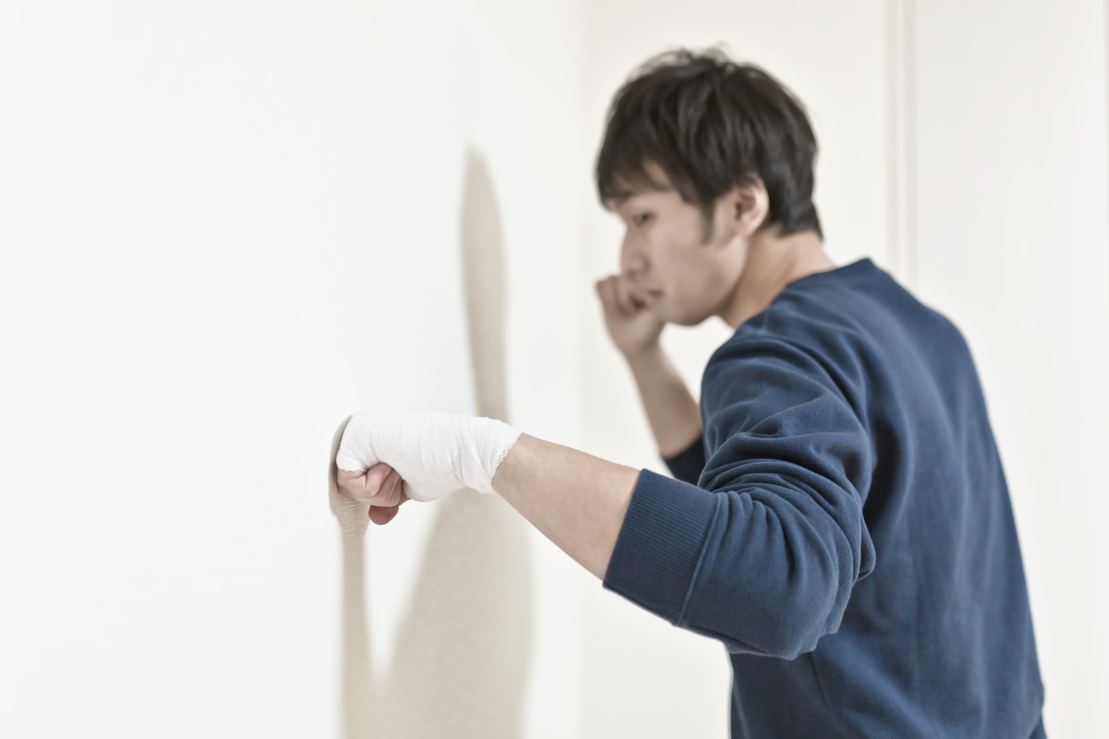 包帯を巻いた右手で壁に拳を突き出す紺色スウェットの男性［モデル：大川竜弥］