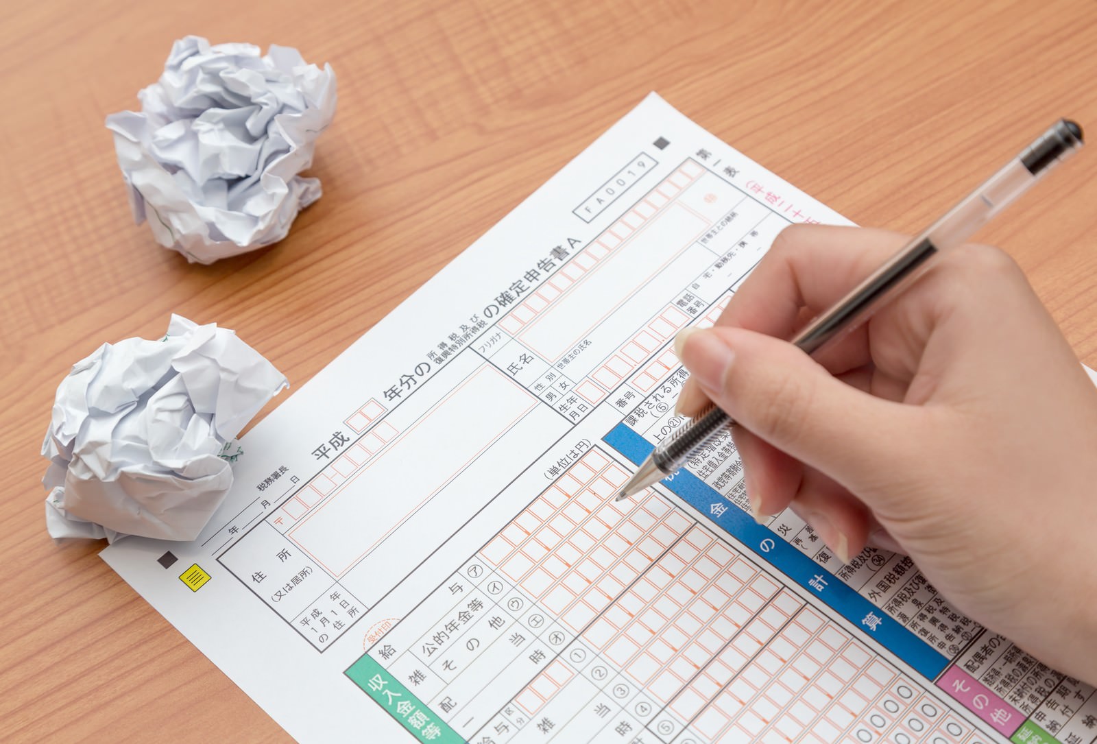 木の机で手がペンを持って確定申告書に記入し横に丸めた白い紙が2つ置かれている