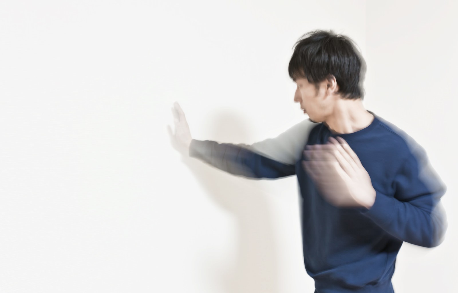 紺色のトップスの男性が白い壁に向かって右腕を伸ばしブレと影が映っている［モデル：大川竜弥］