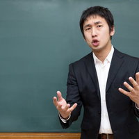 黒板の前で両手を広げて話すスーツの男性の写真