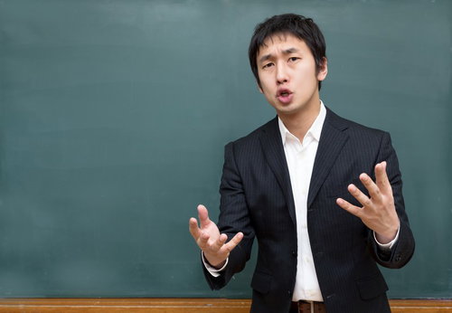 黒板の前で両手を広げて話すスーツの男性