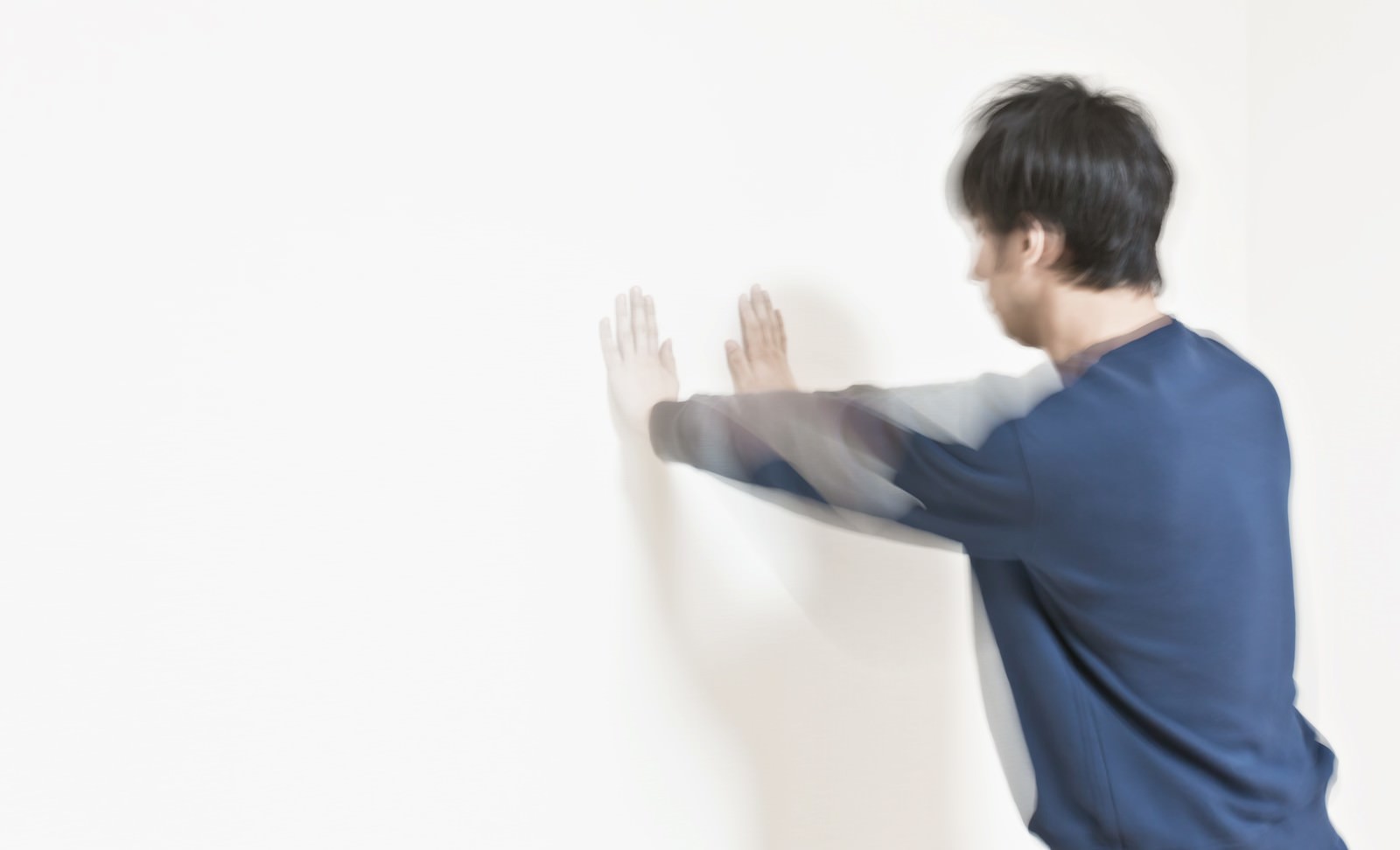 青いシャツの男性が白い壁に両手のひらを押し当てて横向きに立っている［モデル：大川竜弥］