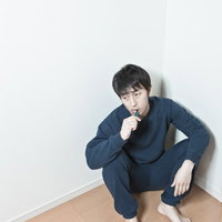 部屋の隅で歯ブラシを口に当てる紺色スウェットの男性の写真