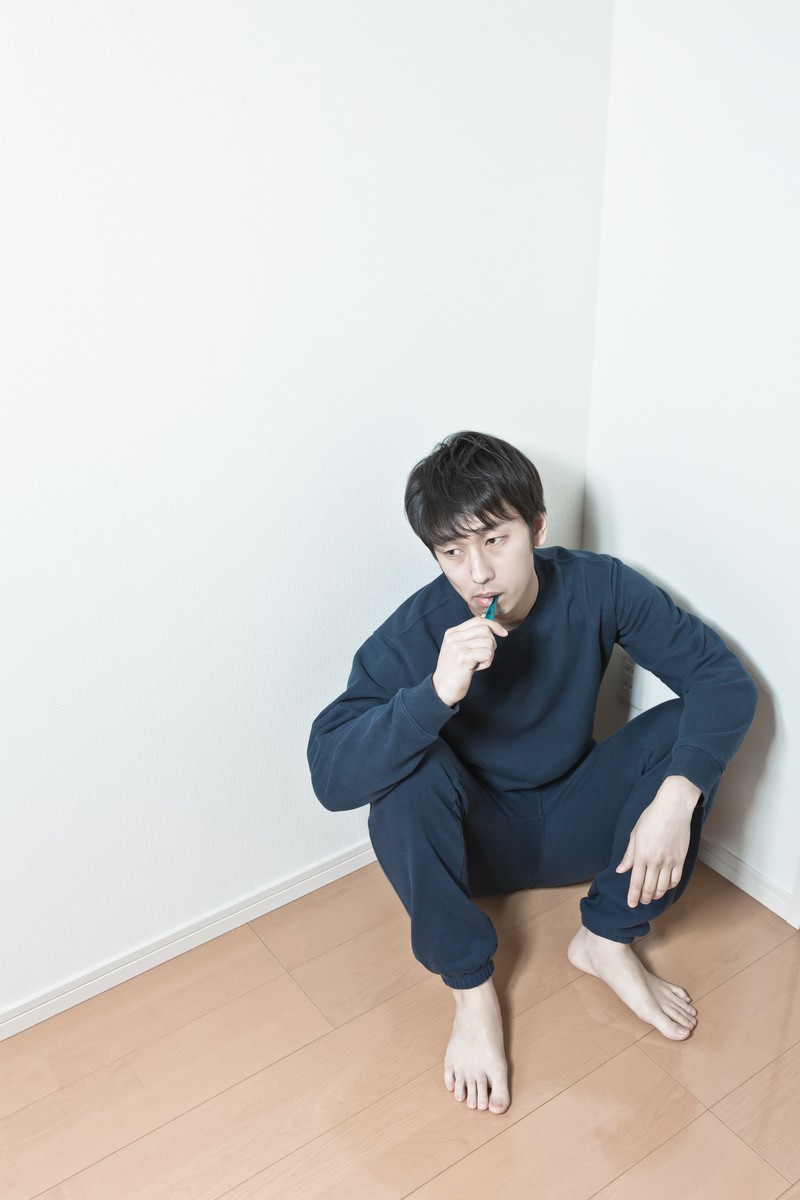 紺色のスウェットの男性が部屋の隅にしゃがんで青い歯ブラシを口に当てている［モデル：大川竜弥］