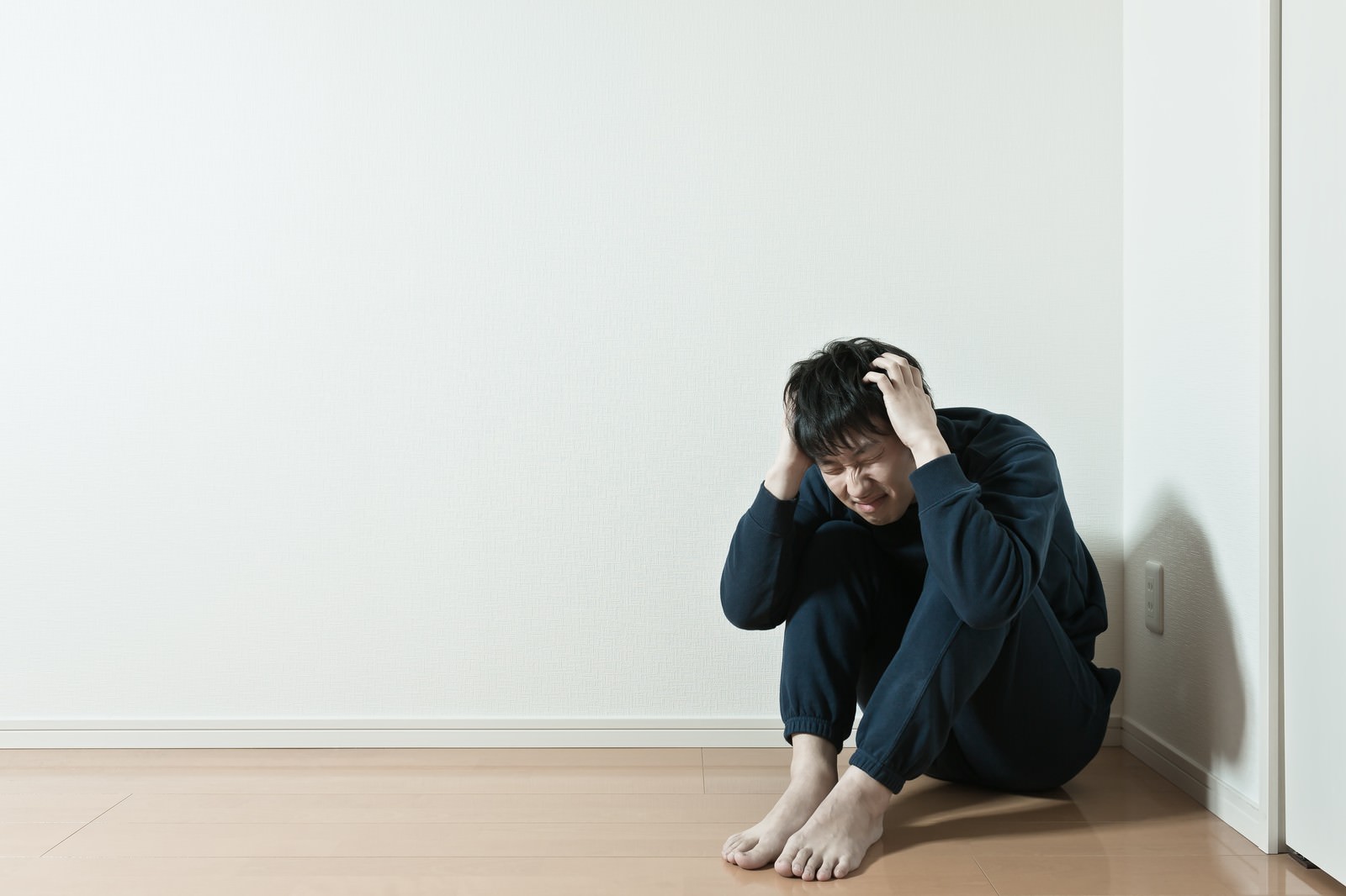 白い壁と木の床の室内で黒い服の男性が膝を抱えて両手で頭を抱えている［モデル：大川竜弥］