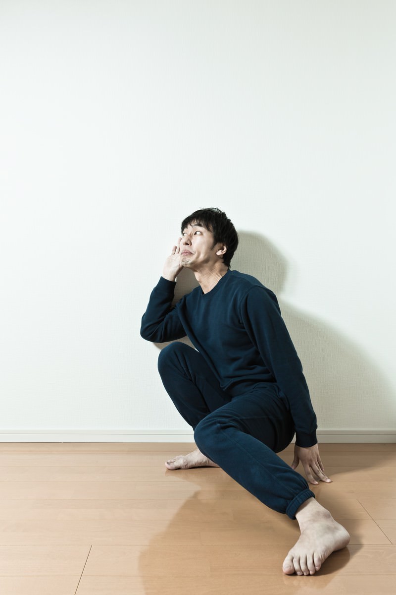 白い壁と木目の床の室内で紺色のスウェットの男性が床に座り左上を見上げている［モデル：大川竜弥］