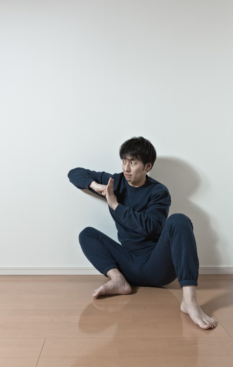 白い壁を背に床に座り、両手を顔の横で構えて横を見る紺色スウェットの男性［モデル：大川竜弥］