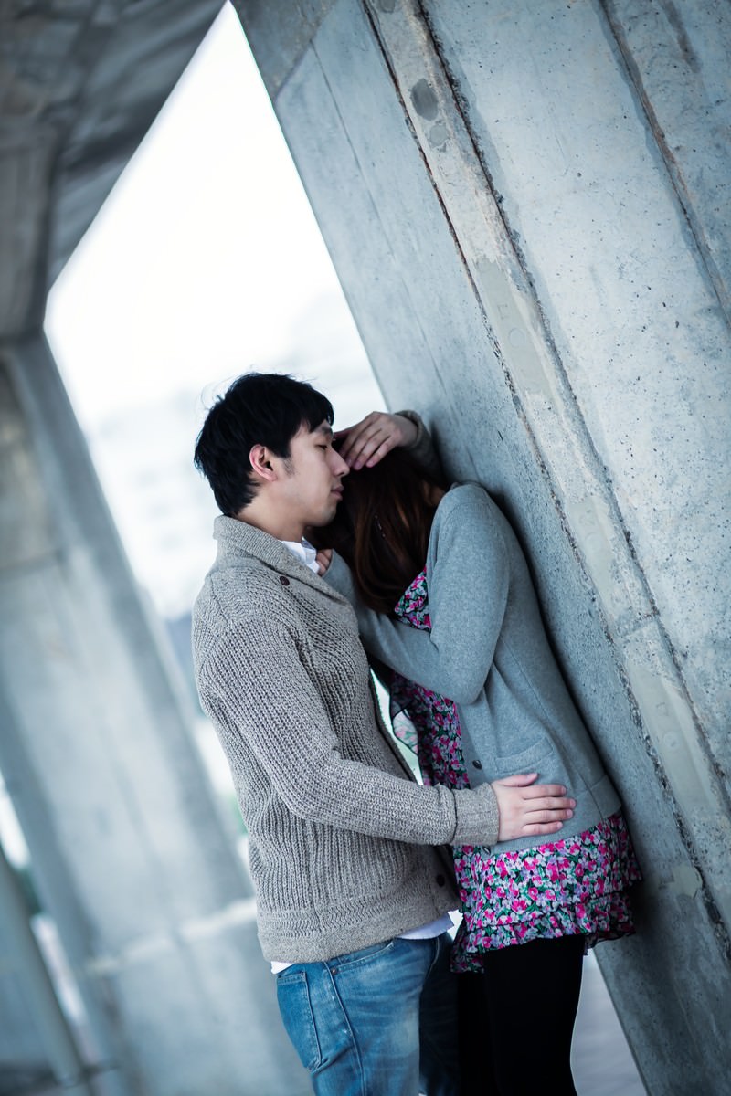 屋外のコンクリート壁の前で茶色のセーターの男性と花柄の女性が寄り添っている［モデル：大川竜弥］