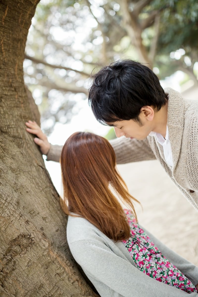 大きな木の幹に花柄の女性とカーディガンの男性が寄り添い下から見上げた構図［モデル：大川竜弥］