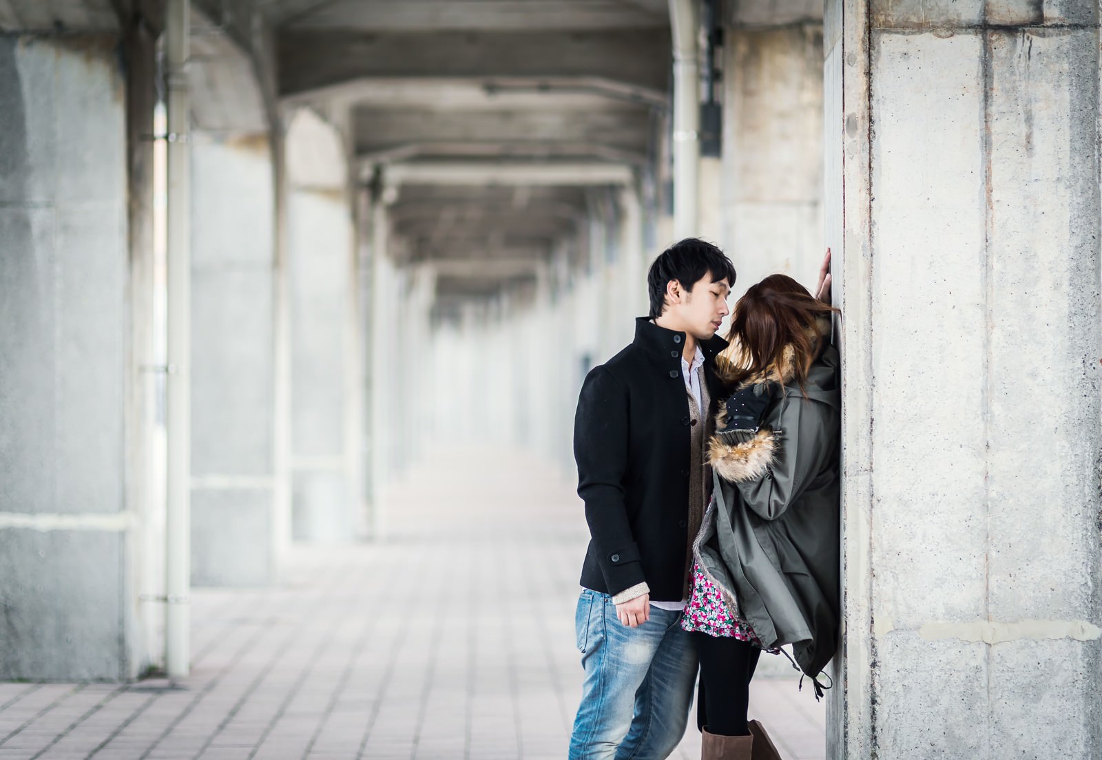 コンクリート柱が並ぶ回廊でジャケットの男性とファーコートの女性が立っている［モデル：大川竜弥］