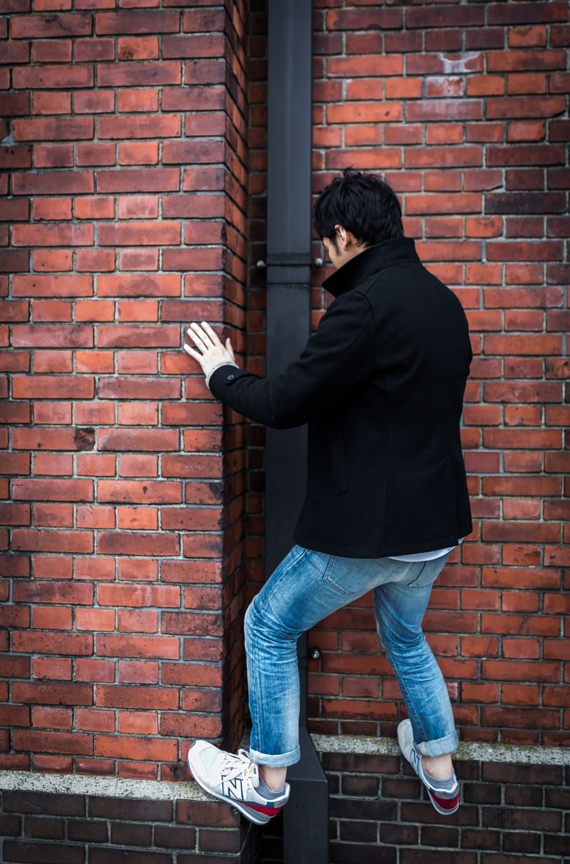 赤レンガの壁に手をついて立つ黒いジャケット姿の男性の後ろ姿［モデル：大川竜弥］