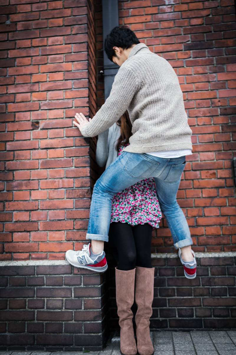 赤レンガの壁でベージュのセーターの男性が壁に手をつき花柄の女性と向かい合っている［モデル：大川竜弥］