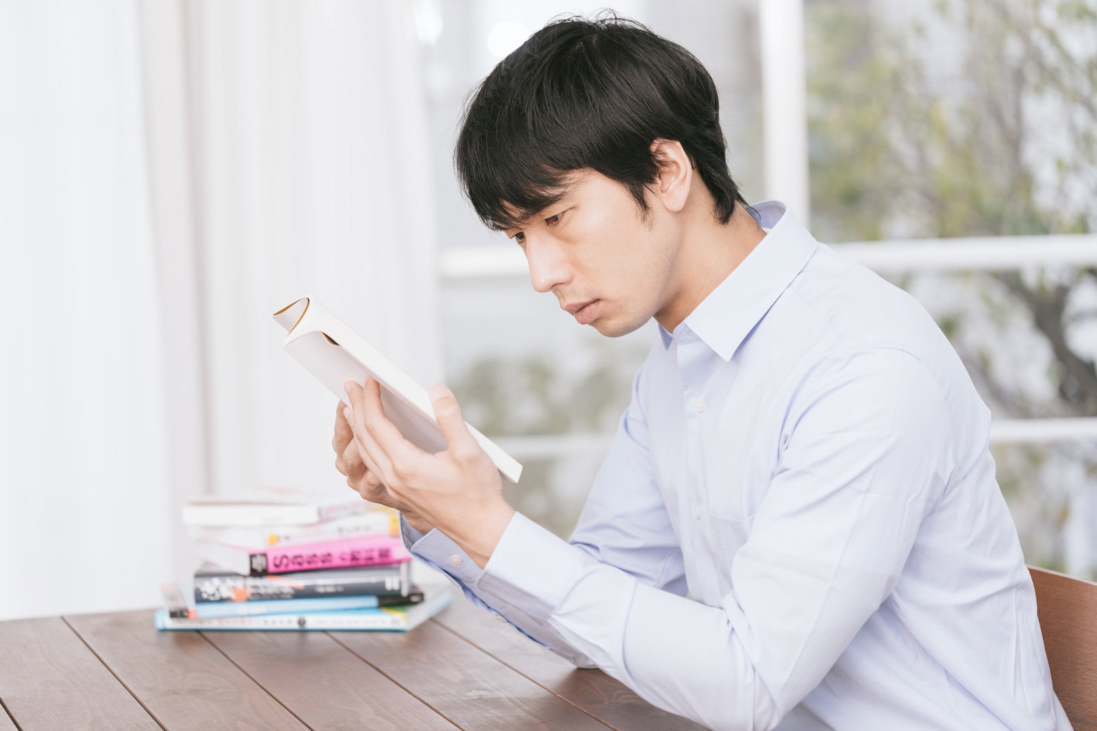 積み重ねた本の前で書籍を見つめて悩む男性［モデル：大川竜弥］