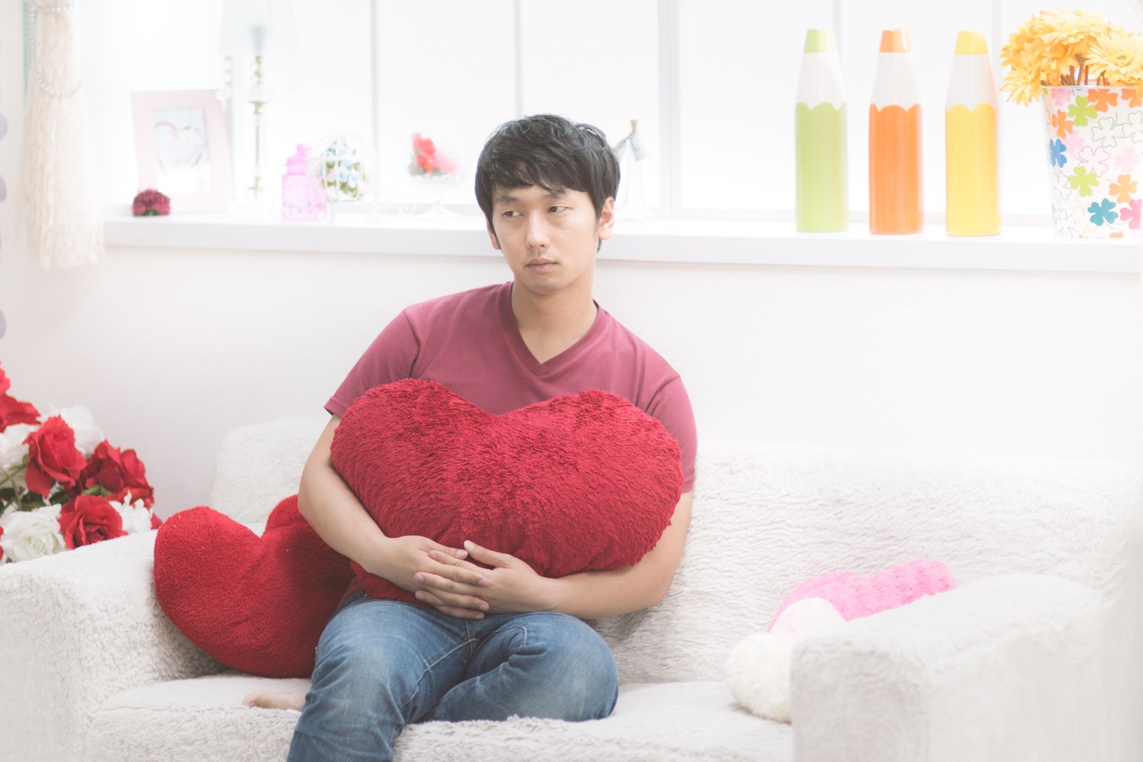 赤いハート型クッションを抱えて悩む男性がソファーに座っている［モデル：大川竜弥］