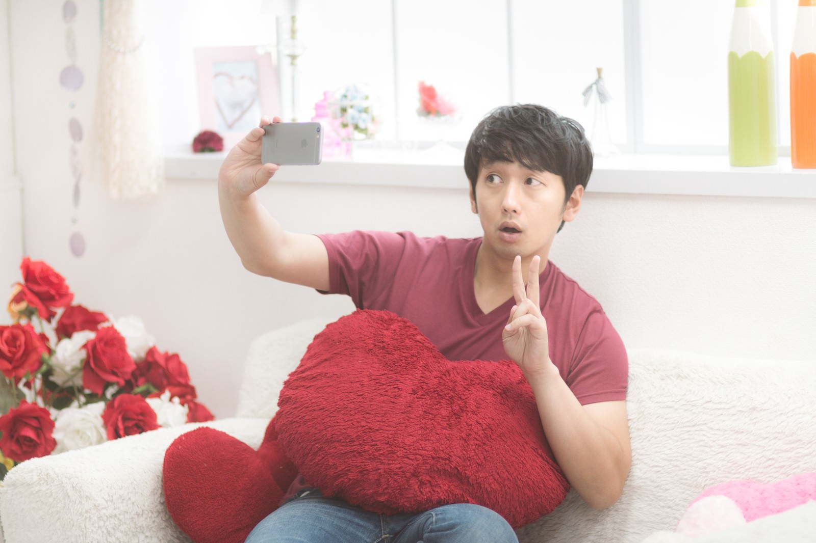 赤いハート型クッションを抱えながらセルフィを撮影する男性［モデル：大川竜弥］