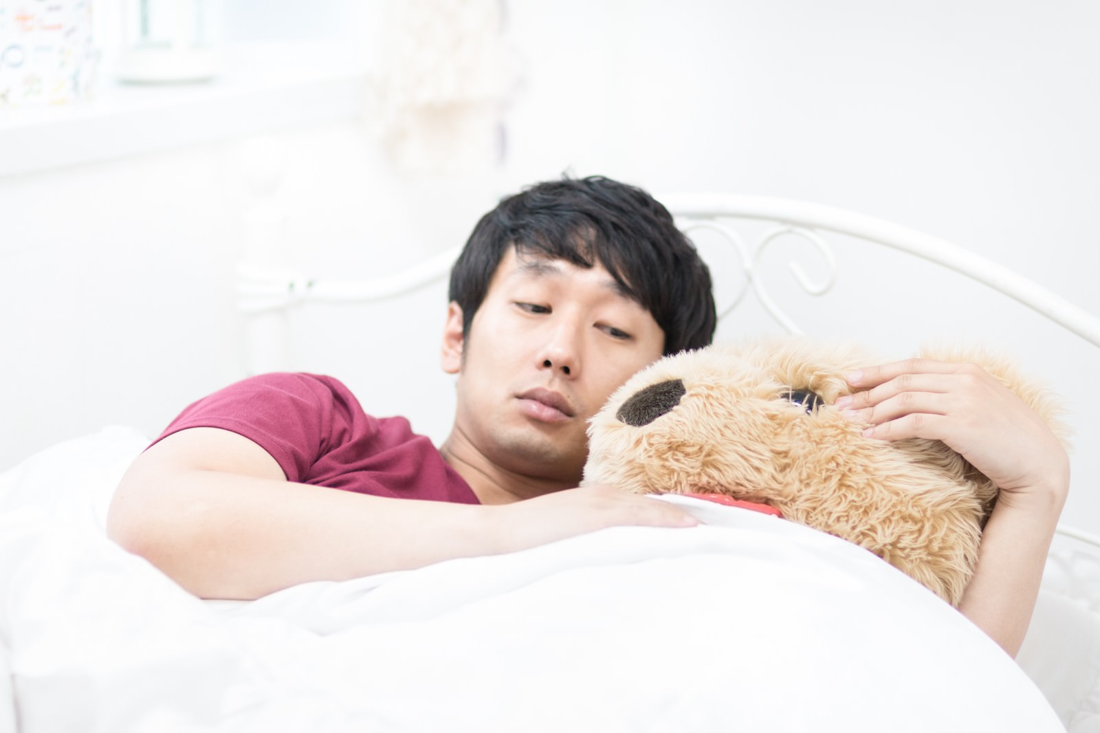 白いベッドの上に横たわり、テディベアを抱きしめる深紅のTシャツ姿の男性［モデル：大川竜弥］