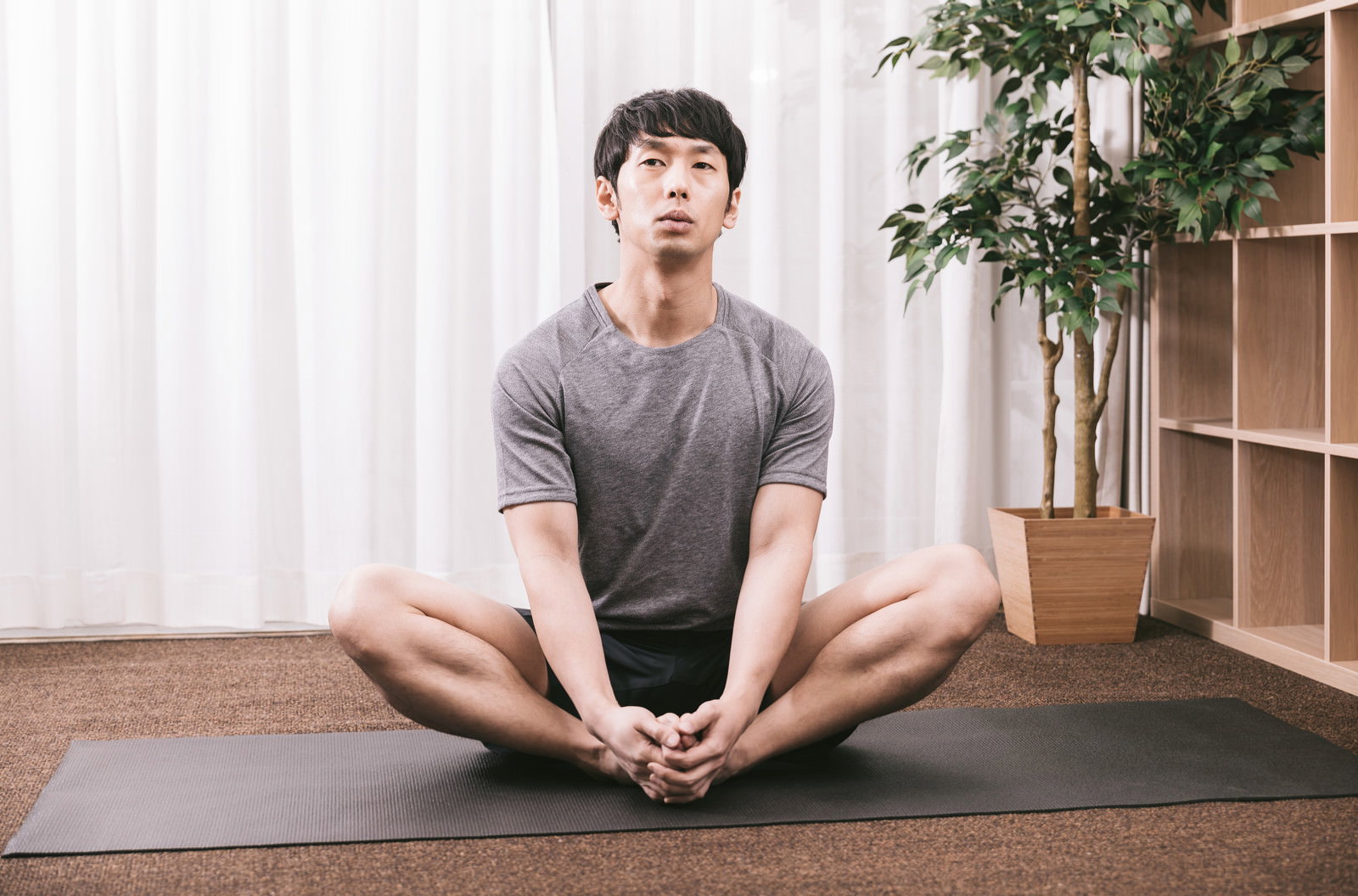マットの上で足を開いて座りながらストレッチの準備をしている男性［モデル：大川竜弥］