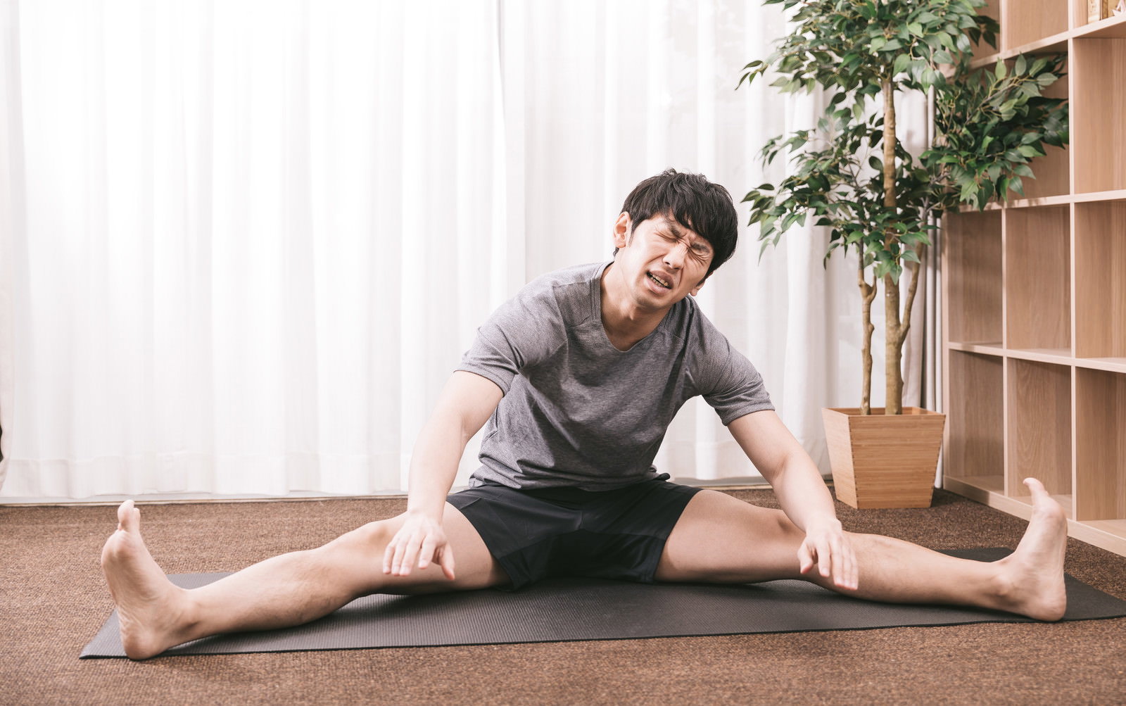 室内でヨガマットの上で開脚ストレッチをするグレーTシャツ姿の男性［モデル：大川竜弥］