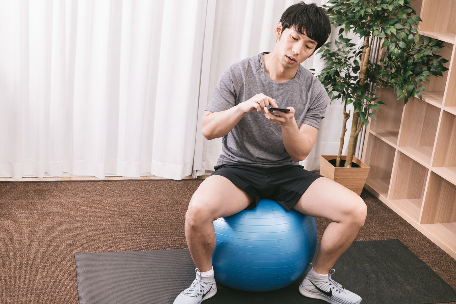 バランスボールに座ってスマートフォンを見る男性［モデル：大川竜弥］