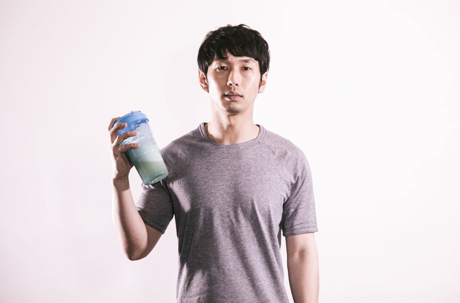 グレーのTシャツを着た男性が青いプロテインシェイカーを持つ［モデル：大川竜弥］