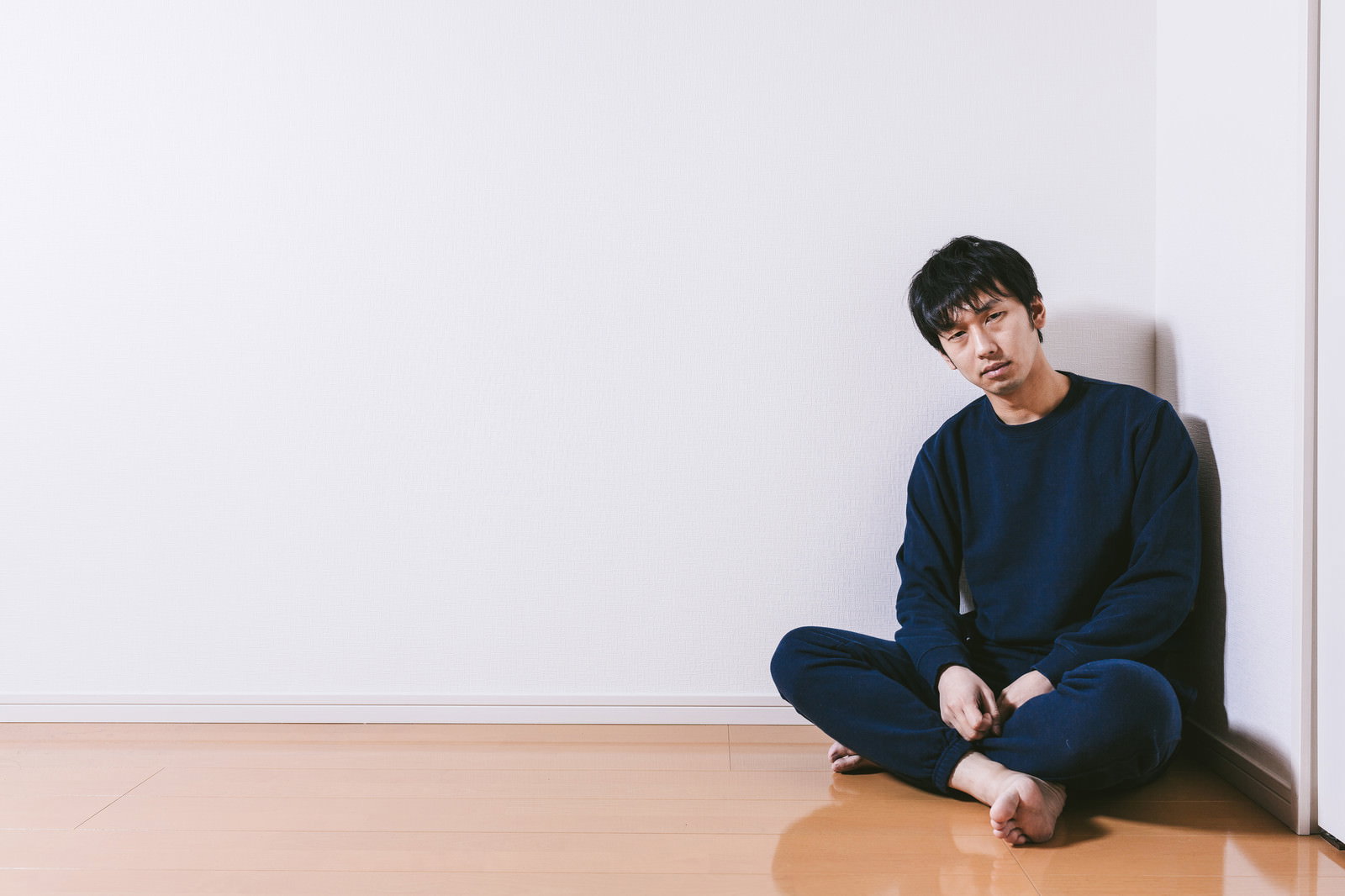 白い壁の部屋の隅でスウェット姿のままあぐらをかいて座り込んでいる男性［モデル：大川竜弥］