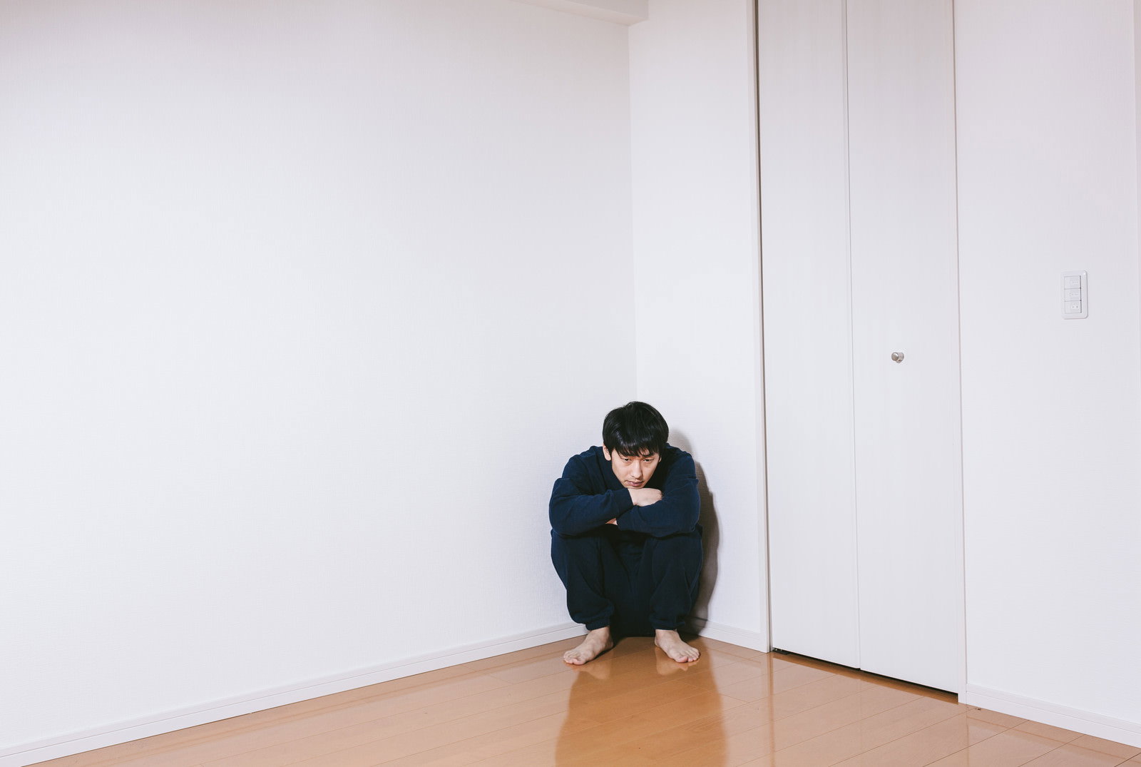 部屋の角で膝を抱えてうつむく男性［モデル：大川竜弥］